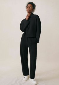 Czarny skrócony sweter z wysokim kołnierzem, zestawiony z dopasowanymi czarnymi spodniami i białymi trampkami, na jasnobeżowym tle.