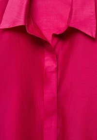 Chemise rose vif à col avec patte de boutonnage cachée, montrant la texture du tissu et les détails des coutures.