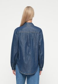 Mørkeblå denimskjorte med spiss krage, puffede lange ermer og en buet kant med knappedetaljer på ermene.