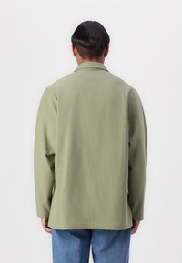 Olive green cotton shirt jacket with long sleeves, a relaxed fit, and a collared neckline, ending below the waist. Classic design details.
