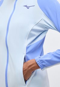 Χέρι στην τσέπη μπουφάν με φερμουάρ σε ανοιχτό μπλε και λεβαντί απόχρωση, με ελαστική μανσέτα και λογότυπο Mizuno στο στήθος.