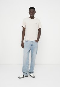 Μπεζ ανοιχτού χρώματος μπλουζάκι με κοντά μανίκια και μικρό λογότυπο, συνδυασμένο με φωτεινό μπλε τζιν σε άνετη γραμμή και μαύρα και λευκά αθλητικά παπούτσια.