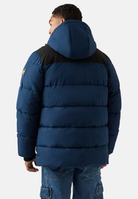 Giacca a vento imbottita blu navy con rinforzo nero sulle spalle, cappuccio oversize, tessuto trapuntato e patch con logo sulla manica sinistra. Abbinata a jeans azzurri.