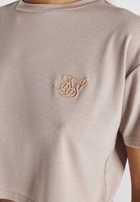 Lyhythihainen, vaalean beige cropped-t-paita, joka on valmistettu hengittävästä kankaasta. Tässä paidassa on tekstuuripinta ja pieni, ruusukultainen brodeerattu logo.