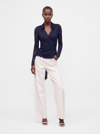 Femme portant un chemisier cache-cœur en dentelle bleu marine à manches longues et un pantalon blanc à jambes larges, debout avec une main dans la poche.