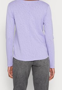 Długo rękawowa koszula w kolorze lawendowym z okrągłym dekoltem i lekko teksturowanym materiałem. Połączenie z szarymi dżinsami, prezentującymi prosty design.