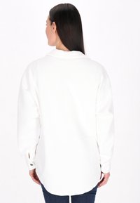 Camicia bianca a maniche lunghe con motivo a spina di pesce testurizzato, vestibilità leggermente oversize e fessure laterali all'orlo, abbinata a jeans scuri.