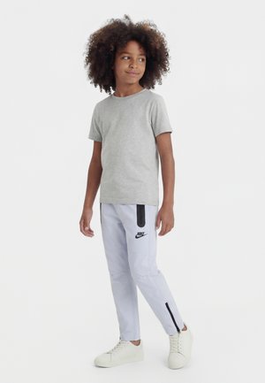 Barn med krøllet hår står iført en grå t-shirt, lysegrå Nike bukser med sorte detaljer og hvide sneakers, og kigger til siden.