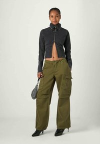 Casaco preto crop com mangas longas e colarinho alto, combinado com calças cargo verde oliva com múltiplos bolsos, e sapatos de salto alto pretos.