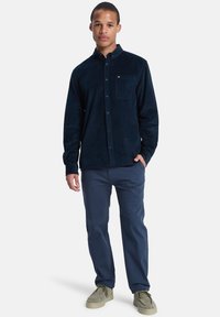 Camisa de pana de color azul oscuro con cuello tipo button-down, un bolsillo en el pecho y mangas largas. Combinada con pantalones azul marino y zapatos beige.