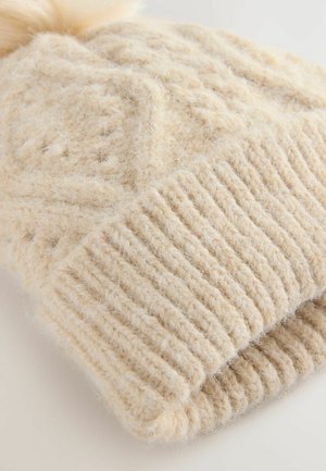 Pletená čepice v jemné béžové barvě, s texturovaným kabelovým vzorem a nadýchaným bambulí na vrchu. Pružný lem na spodní straně dodává detail.