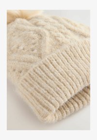 Gorro de punto suave de color crema con un patrón texturizado de cables y puño acanalado. Terminado con un pompón esponjoso en la parte superior.