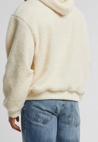 Felpa con cappuccio color crema in pile, con superficie texturizzata, dotata di un ampio cappuccio e polsini a coste. Indossata con jeans di denim azzurri.
