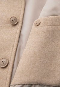 Cappotto di lana marrone chiaro con texture liscia, dotato di due bottoni rotondi e una tasca piegata. Interno foderato di un colore beige chiaro.