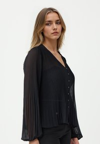 Schwarze plissierte Bluse mit V-Ausschnitt, langen Ärmeln und Knopfverschluss. Hergestellt aus durchsichtigem Stoff mit weicher Textur und lockerer Passform.