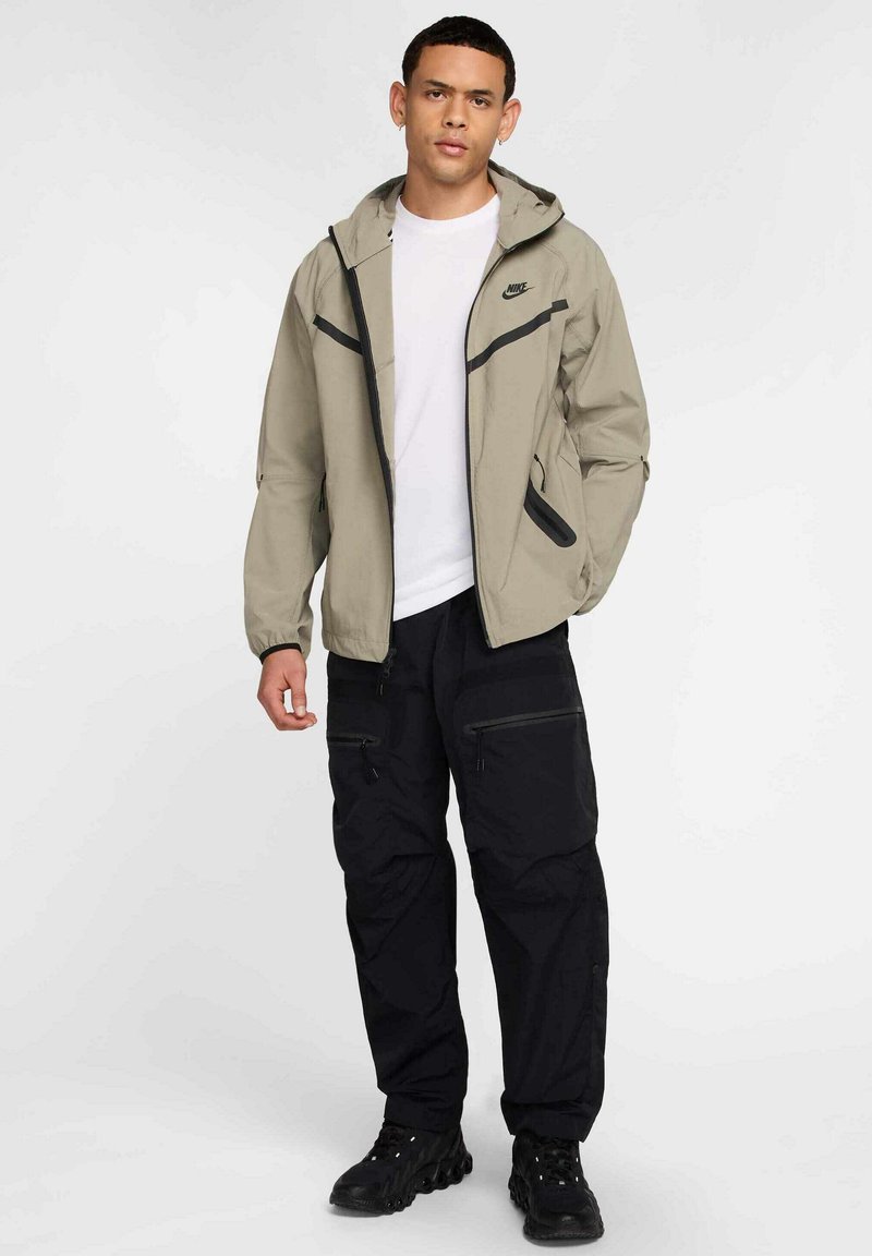 Lätt beige jacka med svarta accenter, dragkedja fram, två sidofickor, tillsammans med svarta cargobyxor och svarta skor.