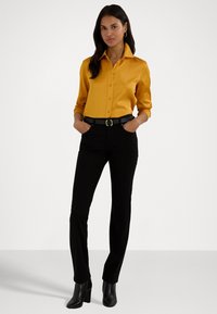 Camisa abotoada de satin mostarda com mangas longas, combinada com calças justas pretas e um cinto preto. Botins de salto pretos completam o visual.