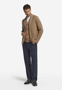 Beige stickad cardigan med sjalkrage över en beige väst, kombinerad med marinblå byxor och svarta skor. Cardiganen har en avslappnad passform och knappar.