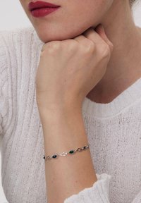 Srebrna bransoletka z owalnymi niebieskimi i zielonymi kamieniami, połączona z łańcuchowym wzorem; noszona na nadgarstku w białym, teksturowanym swetrze.