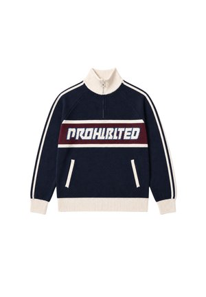Mand iført en marineblå sweater med hvide striber og ordet "PROHBLITE" i store hvide og blå bogstaver på tværs af brystet.