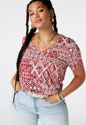 Vrouw met lang gevlochten haar die een rood-wit shirt met V-hals en patroon draagt, lichtblauwe spijkerbroek, oorringen, gelaagde kettingen en armbanden.