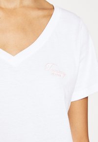Bílý bavlněný tričko s designem do V. Obsahuje malé, růžové vyšívané logo "Tommy Jeans" na levé hrudi. Krátké rukávy.