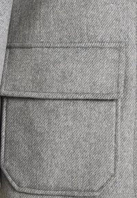 Texture de laine grise avec un motif chevrons diagonal. Comprend une grande poche plaquée carrée aux bords arrondis. Détails de couture soignés visibles.