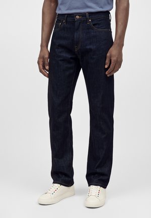 Homme portant un jean bleu foncé avec des surpiqûres orange et des baskets blanches avec des œillets colorés sur un fond gris uni.
