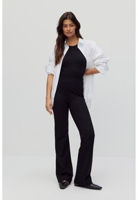 Zwarte fitted jumpsuit met wijde pijpen, gedragen onder een oversized witte blouse met knopen. Glad stof, minimalistisch ontwerp en geen zichtbare patronen.