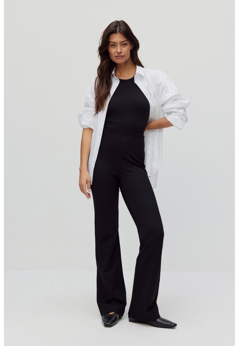 Zwarte fitted jumpsuit met wijde pijpen, gedragen onder een oversized witte blouse met knopen. Glad stof, minimalistisch ontwerp en geen zichtbare patronen.