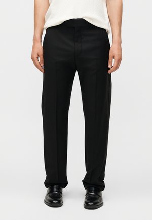 Férfi fekete egyenes szárú nadrágot, fekete bőr cipőt és fehér ujjatlan felsőt visel, egyszínű háttér előtt áll.