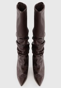 Botas altas, sueltas, de cuero texturizado marrón oscuro con puntas puntiagudas, expuestas de pie sobre un fondo blanco liso.