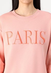 Sudadera rosa con "PARÍS" en letras bordadas elevadas en un tono a juego. Textura suave y cuello de tripulación.
