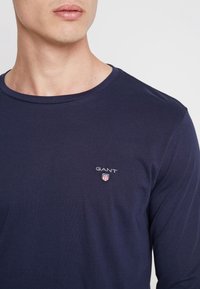 T-shirt a maniche lunghe blu navy realizzato in morbido cotone. Presenta un collo rotondo e un piccolo logo ricamato sul petto con un design a scudo.