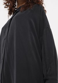 Schwarze, oversized Bluse aus weichem, leichtem Stoff. Verfügt über einen Kragen, eine Knopfleiste und lange Ärmel mit lockeren Manschetten.