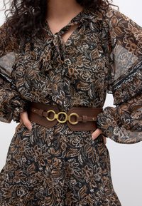 Blouse à motifs floraux aux tons marron et crème, avec des manches amples et un col en forme de goutte, assortie d'une ceinture marron avec des éléments en métal doré.