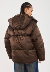 Giacca marrone imbottita con cappuccio, caratterizzata da una finitura lucida e cuciture orizzontali. Le maniche sono elasticizzate per garantire comfort.
