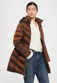Vrouw in een bruine gewatteerde jas met capuchon over een witte gebreide trui, gecombineerd met blauwe jeans, staande tegen een witte achtergrond.