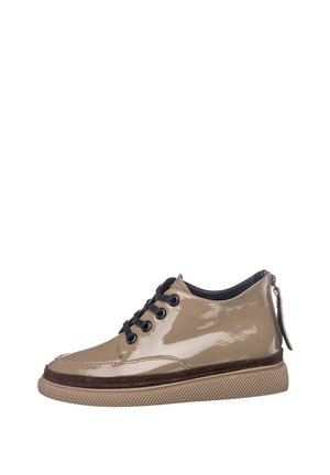 Personne portant un jean bleu clair et des chaussures lacées taupe brillantes avec des semelles épaisses, debout avec un pied légèrement levé sur un fond clair.