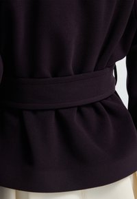 Mörklila jacka med bälte i midjan och mjukt, texturerat tyg. Fållad detaljering på baksidan ger designen djup.