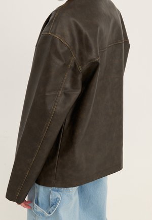 Personne portant une veste en cuir marron foncé et un pantalon en jean bleu clair, vue de dos et de côté sur un fond uni.