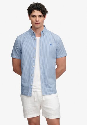Hombre con camisa de manga corta con rayas verticales azul claro y blancas sobre una camiseta blanca y pantalones cortos blancos, con las manos detrás de la espalda.
