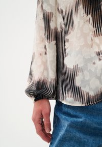 Prosojna bluzica z črnimi, belimi in taupe cvetličnimi vzorci, dolgi rokavi z elastiko na manšetah in pleated teksturo. V kombinaciji z jeansom.