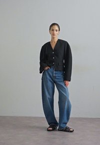 Černý strukturovaný blazer s V-výstřihem a zapínáním na knoflíky, spárovaný s volnými modrými džíny a sandály na platformě s černými hroty.