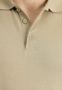 Beżowa koszulka polo wykonana z fakturowanej tkaniny, z kołnierzykiem, trzema guzikami i dyskretną kieszenią na piersi.