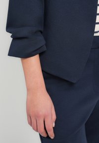 Μανίκι μπλέ σκούρου σακακιού με ρεβέρ, συνδυασμένο με παντελόνι σε ίδια απόχρωση. Το ύφασμα φαίνεται υφασμάτινο με ελαφριά ελαστικότητα.