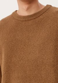 Hnědý pletený svetr s žebrovaným kulatým výstřihem, jemnou texturou a volným střihem. Materiál vypadá teple a pohodlně.