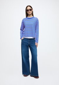 Pruhovaná modrobiela blúzka s dlhým rukávom, kombinovaná s modrými džínsami so širokými nohavicami. Modelka má na sebe aj tmavé obdĺžnikové slnečné okuliare.