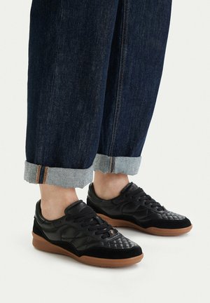 Stopy w czarnych pikowanych trampkach z brązowymi podeszwami, w połączeniu z ciemnoniebieskimi podwiniętymi dżinsami na białym tle.