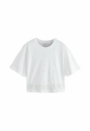 Kvinde med langt blond hår iført en hvid løs top og shorts står barfodet på våd sand nær rolige oceanbølger.